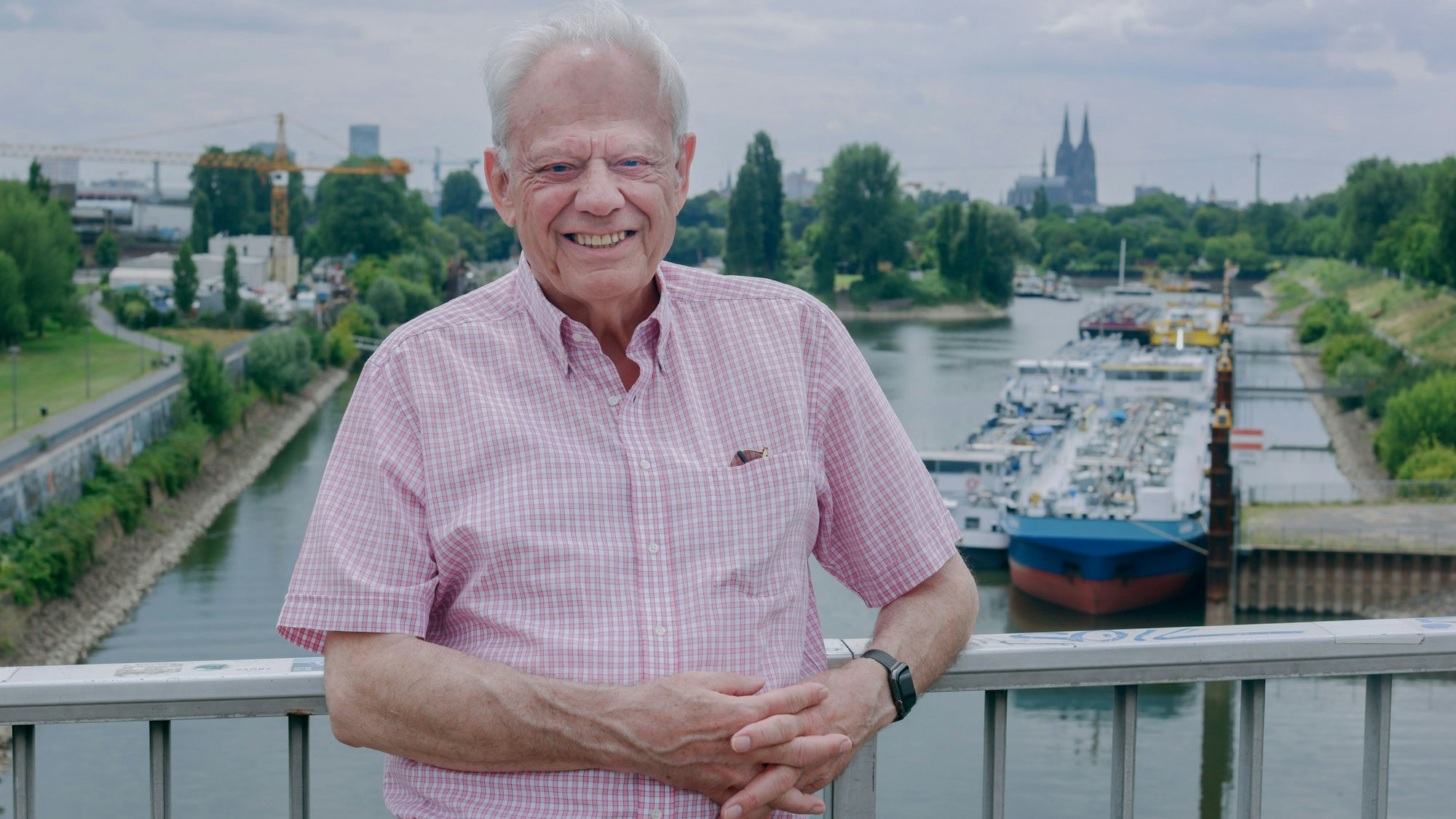Ein Mann im rosa-weißen Karohemd steht auf einer Brücke. Im Hintergrund sind das Mülheimer Hafenbecken und der Dom zu sehen.