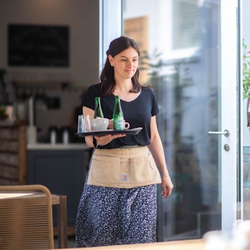 Eine junge Frau läuft mit einem Tablett, auf dem Kaffeetassen und Wasserflaschen stehen, zu einem Tisch.