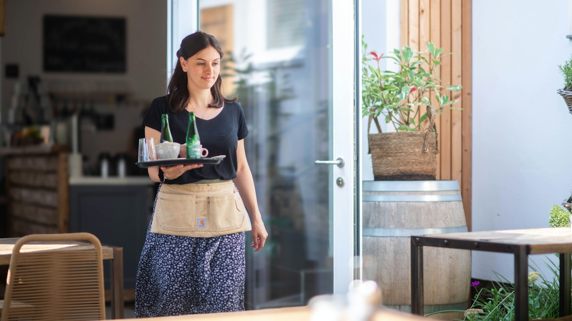 Eine junge Frau läuft mit einem Tablett, auf dem Kaffeetassen und Wasserflaschen stehen, zu einem Tisch.