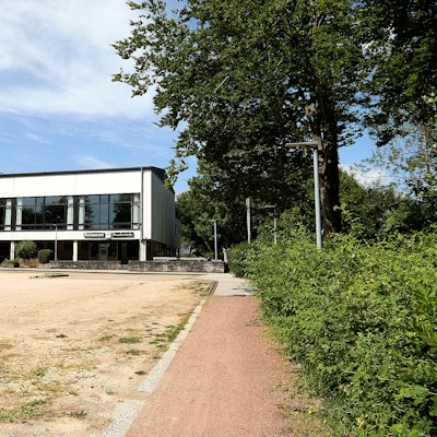 Im Vordergrund ist ein geschotterter Parkplatz, im Hintergrind die Grenzlandhalle in Hellenthal.