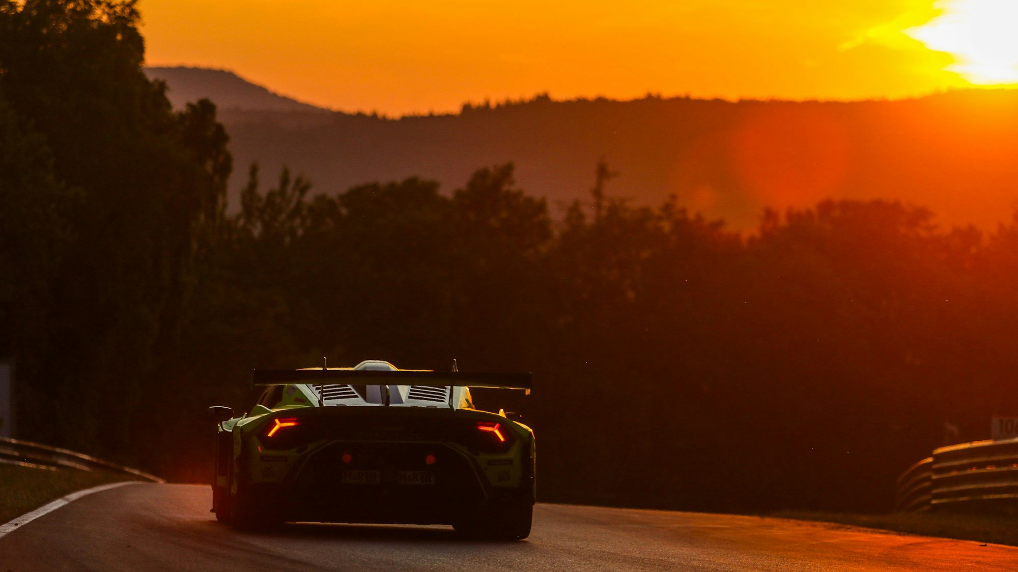 Das Bild zeigt einen Rennwagen, der auf dem Nürburgring fährt. Im Hintergrund geht die Sonne unter.