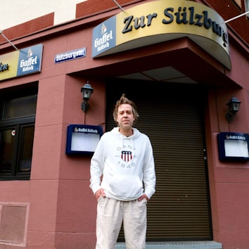 Ein Mann steht vor einer Eckkneipe mit heruntergelassenen Rollläden.