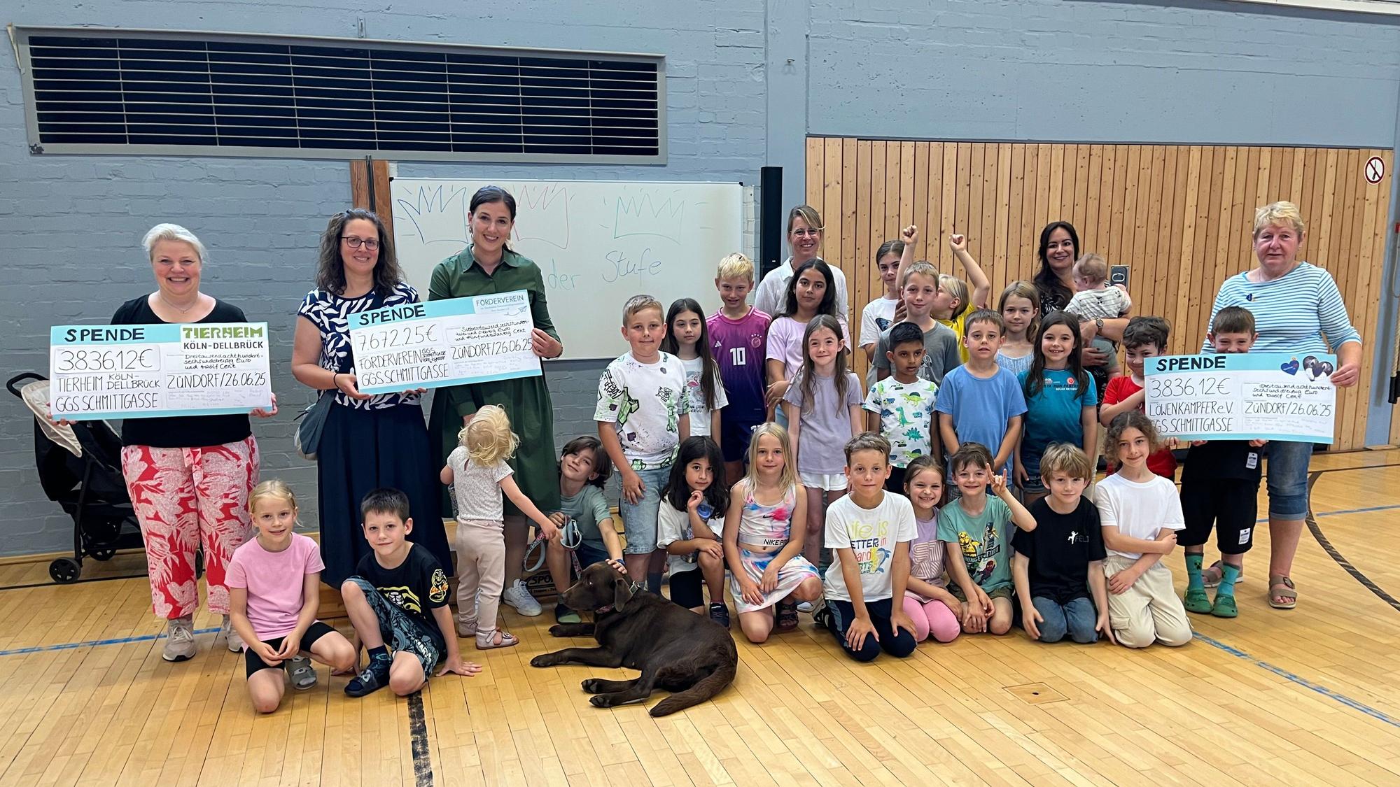 Viele Kinder hocken in einer Turnhalle, ein Hund ist auch dabei. Vier Frauen halten großformatige Spenden-Schecks in die Höhe.