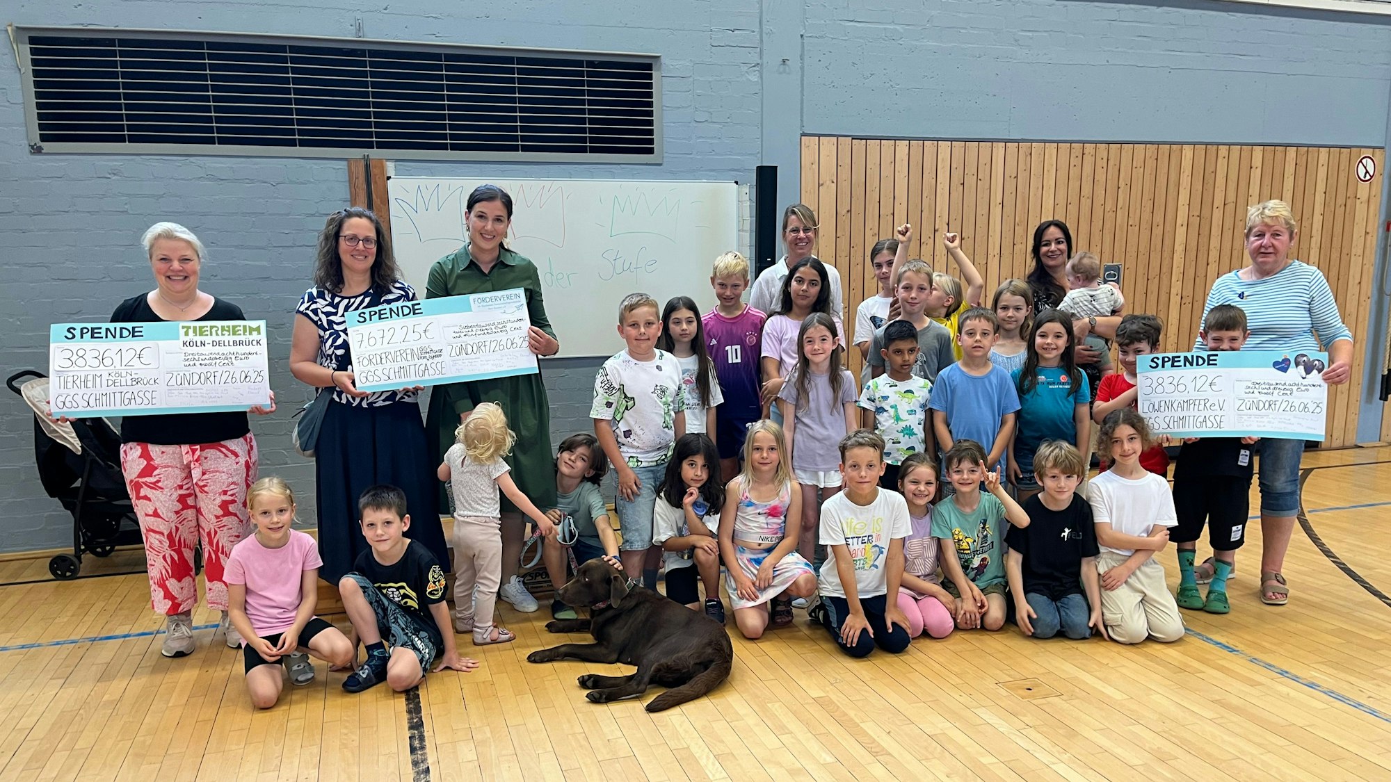 Viele Kinder hocken in einer Turnhalle, ein Hund ist auch dabei. Vier Frauen halten großformatige Spenden-Schecks in die Höhe.