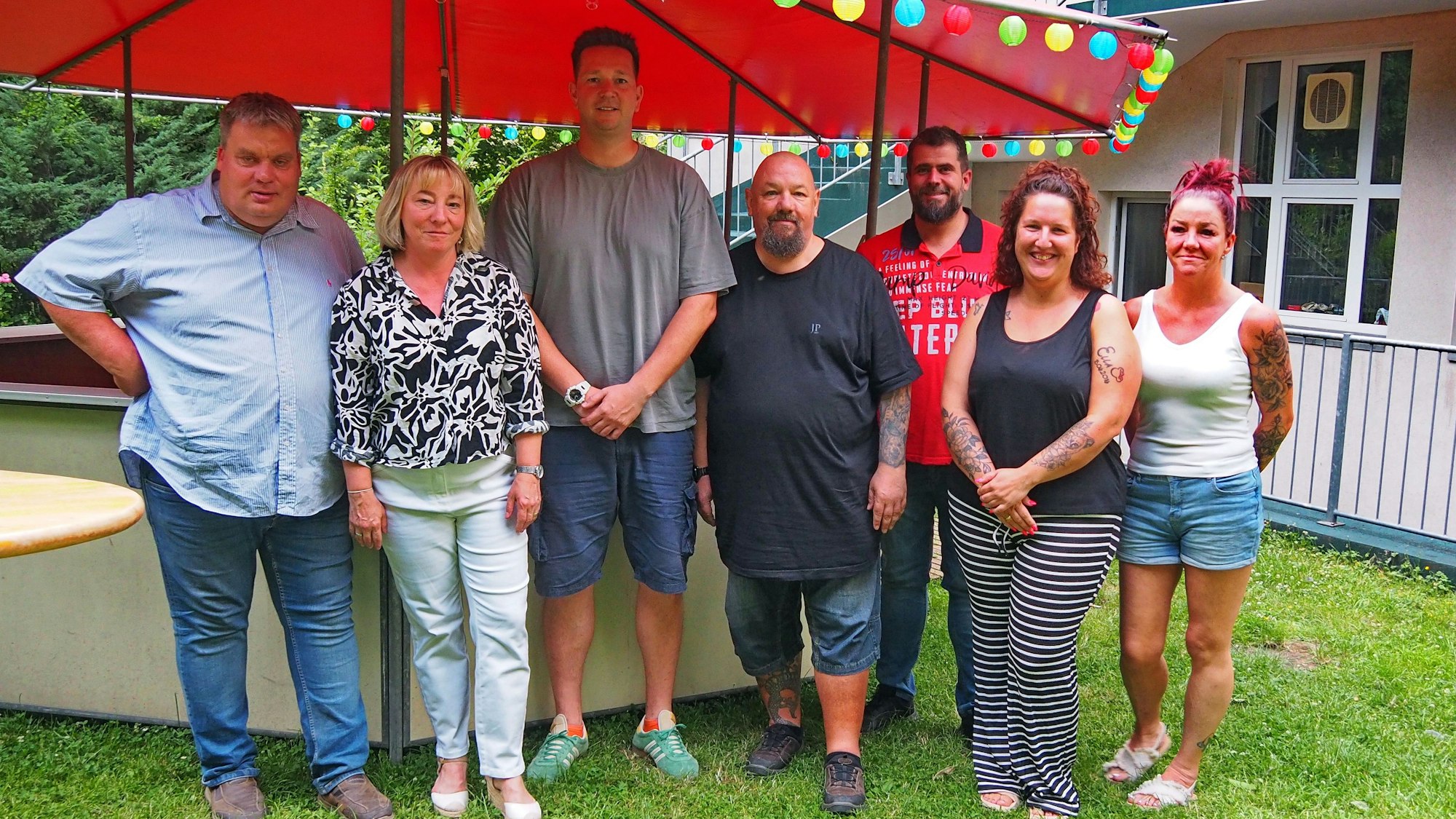 Sieben Menschen stehen nebeneinander vor einem Pavillon mit rotem Dach.