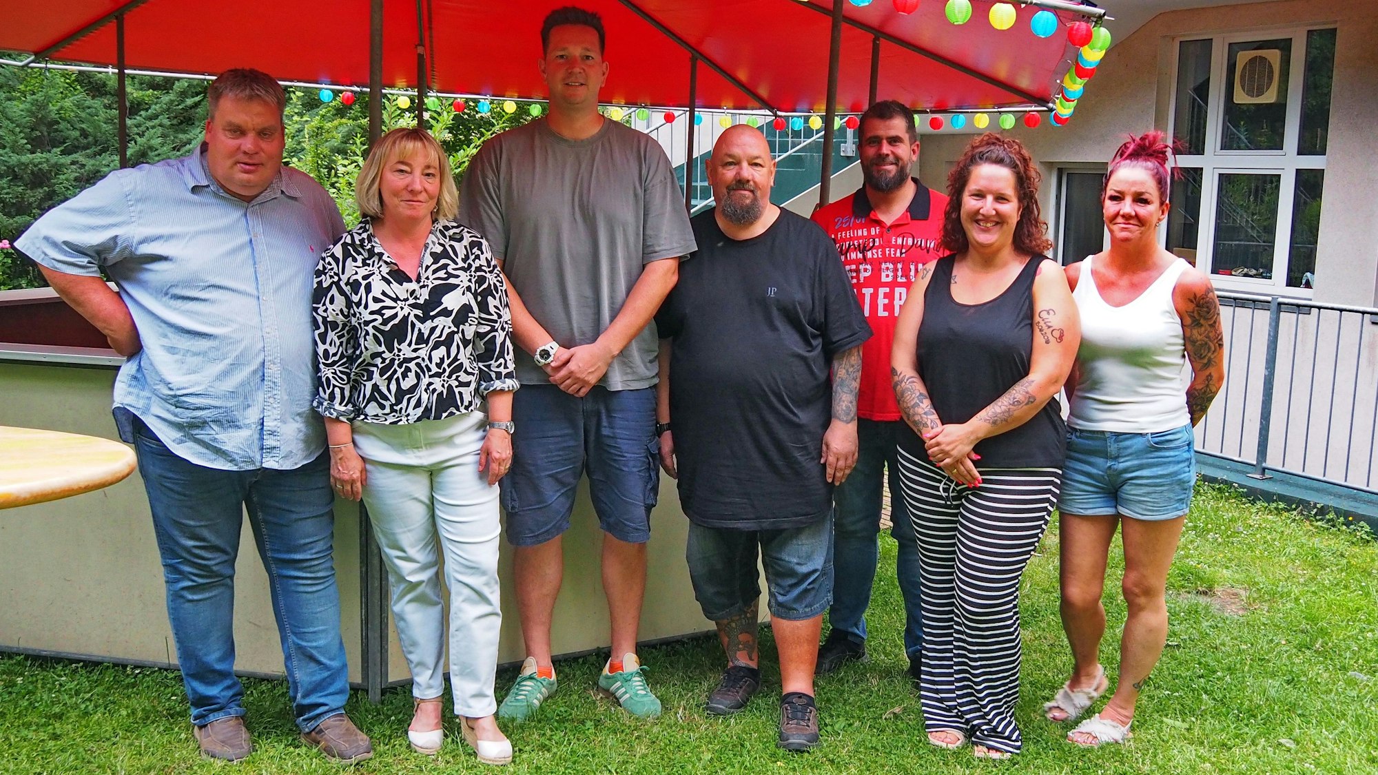 Sieben Menschen stehen vor einem Bierstand mit bunter Lichterkette.