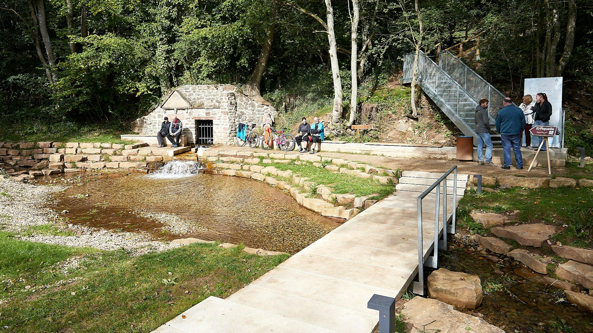 Blick auf eine Brücke und die Erftquelle, neben der einige Menschen sitzen.