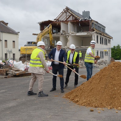 Vier Männer mit Schutzhelmen werfen mit Spaten Erde in die Luft. Im Hintergrund bricht ein Bagger ein Gebäude ab.