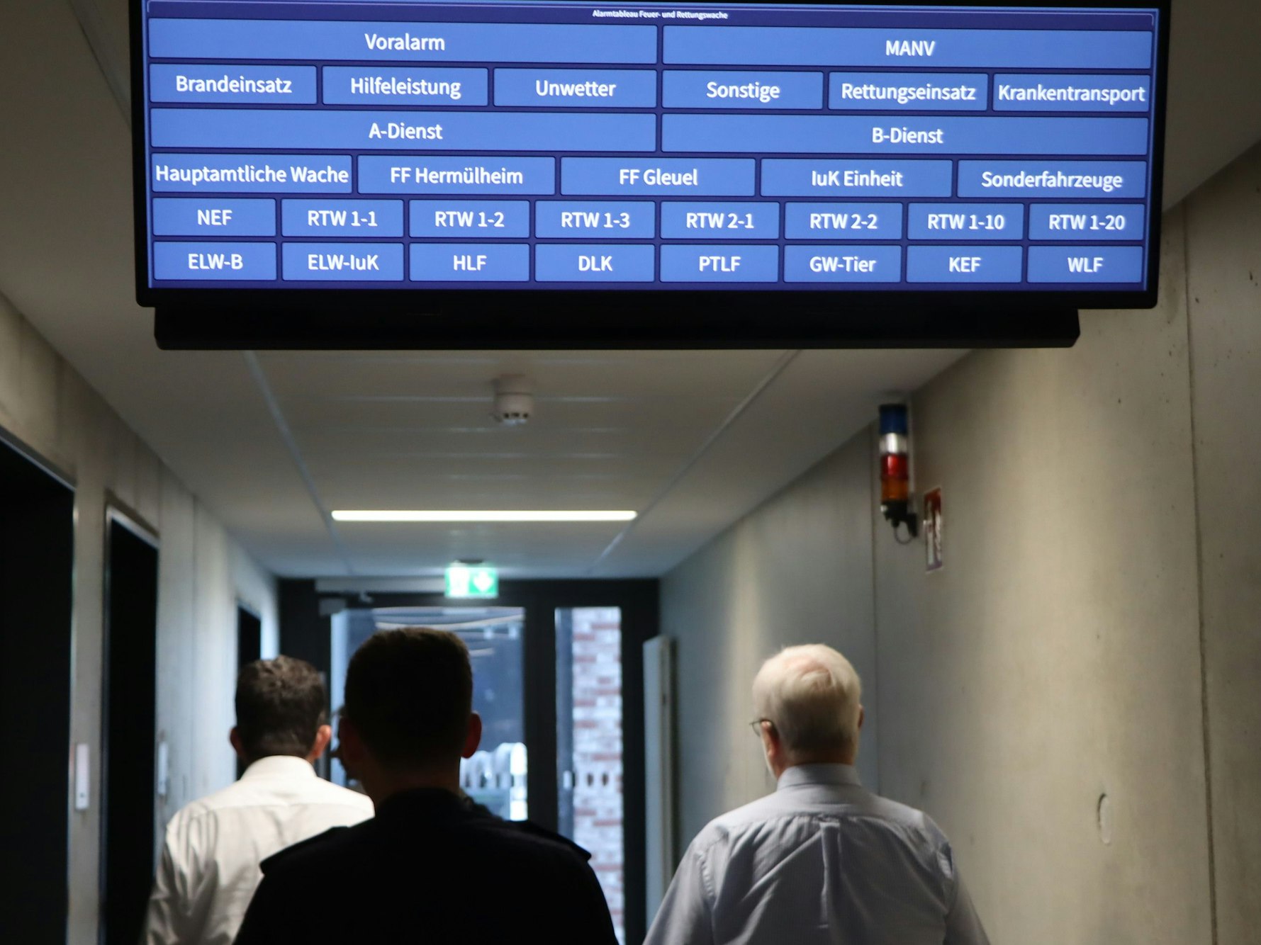 Das Bild zeigt eine digitale Anzeigetafel und drei Feuerwehrleute in einem Gang.