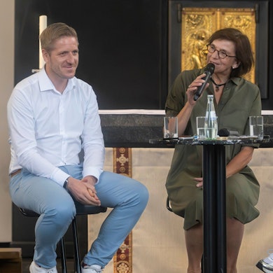 Das Bild zeigt einen Moment der Talkrunde. Marianne Komp hält ein Mikrofon in der Hand, die beiden Landratskandidaten sitzen auf Stühlen und lauschen.
