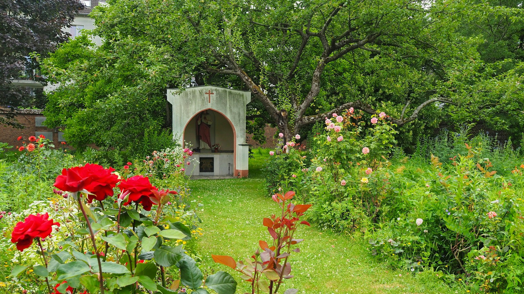 In einem Garten mit Rasenflächen und blühenden Rosen befindet sich auch eine kleine offene Kapelle zum Beten.