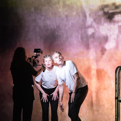 Patrycia Ziolkowska und Sebastian Blomberg stehen leicht gebückt in der Opernkulisse. Sie tragen weiße T-Shirts und schwarze Hosen. Eine Frau filmt sie.