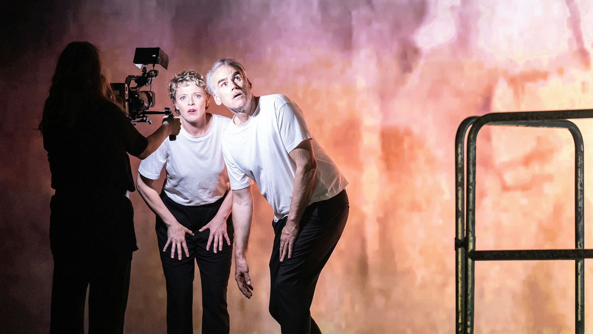 Patrycia Ziolkowska und Sebastian Blomberg stehen leicht gebückt in der Opernkulisse. Sie tragen weiße T-Shirts und schwarze Hosen. Eine Frau filmt sie.