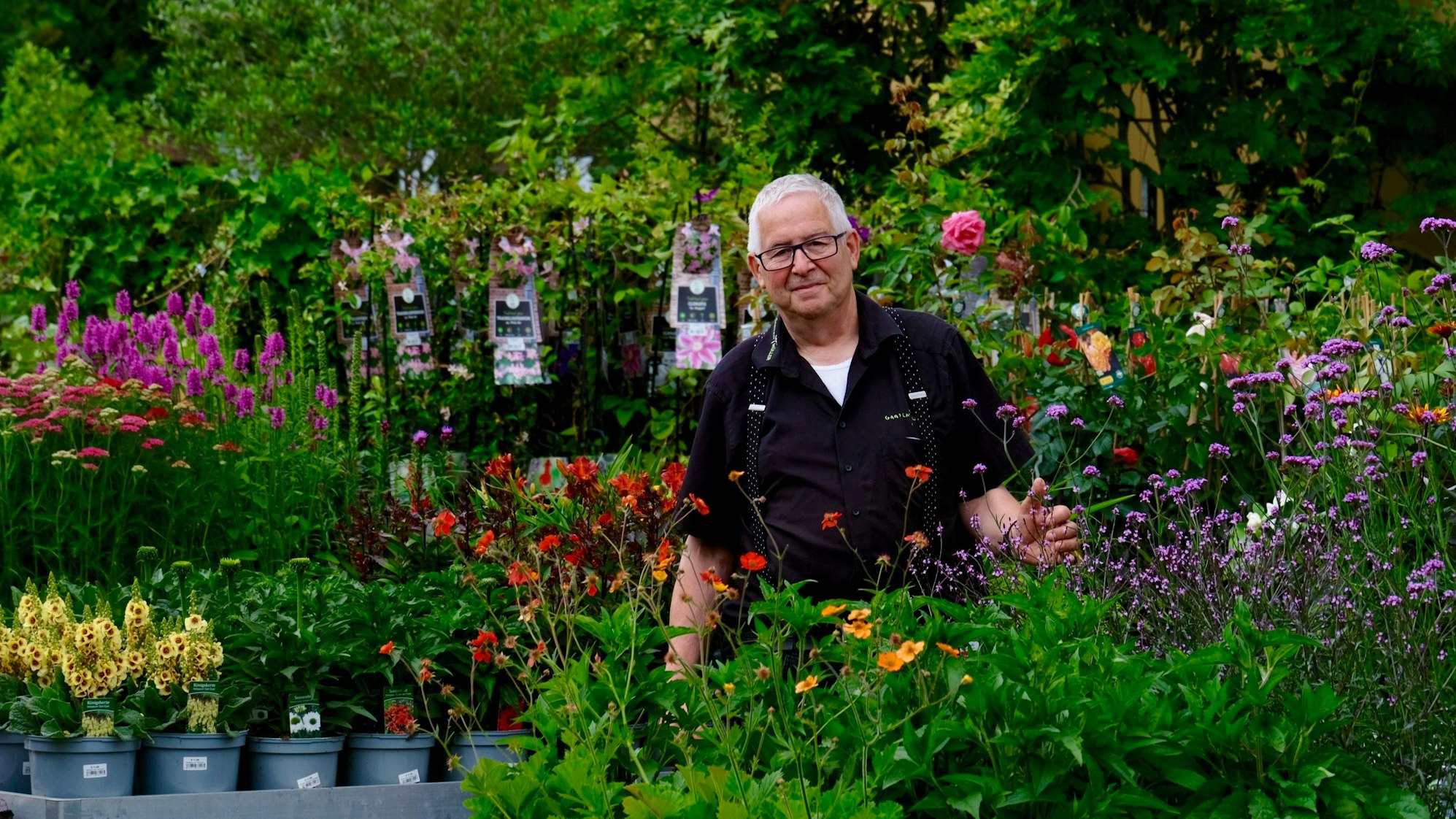 Ein Mann mit kurzem weißem Haar und Brille steht inmitten von Pflanzen.