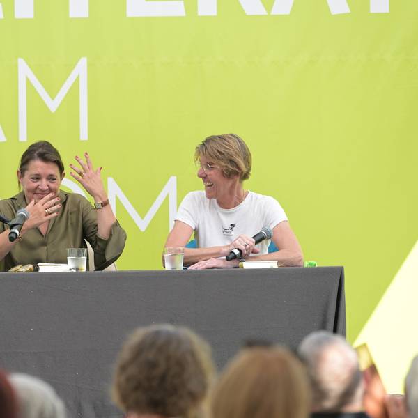 Zwei Frauen sitzen auf einer Bühne, hinter ihnen eine grasgrüne Wand.
