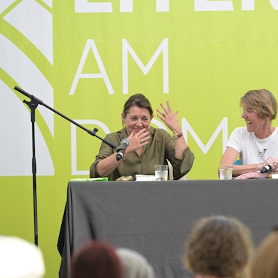 Zwei Frauen sitzen auf einer Bühne, hinter ihnen eine grasgrüne Wand.
