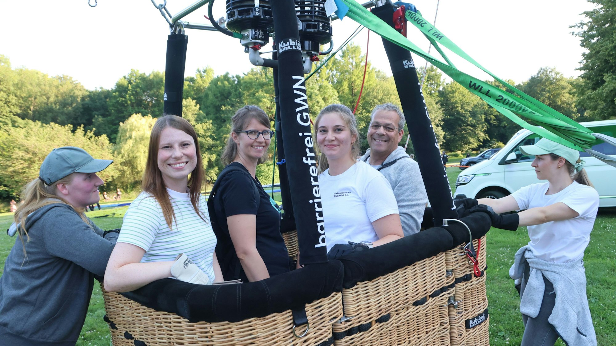 Vier Personen, ein Mann und drei Frauen stehen im Korb eines Heißluftballons. Zwei Frauen an den Seiten drücken den Korb von außen runter.