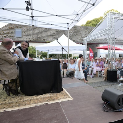 Zwei Männer sitzen auf einer Bühne, davor in einem Innenhof das Publikum unter einem Sonnendach.