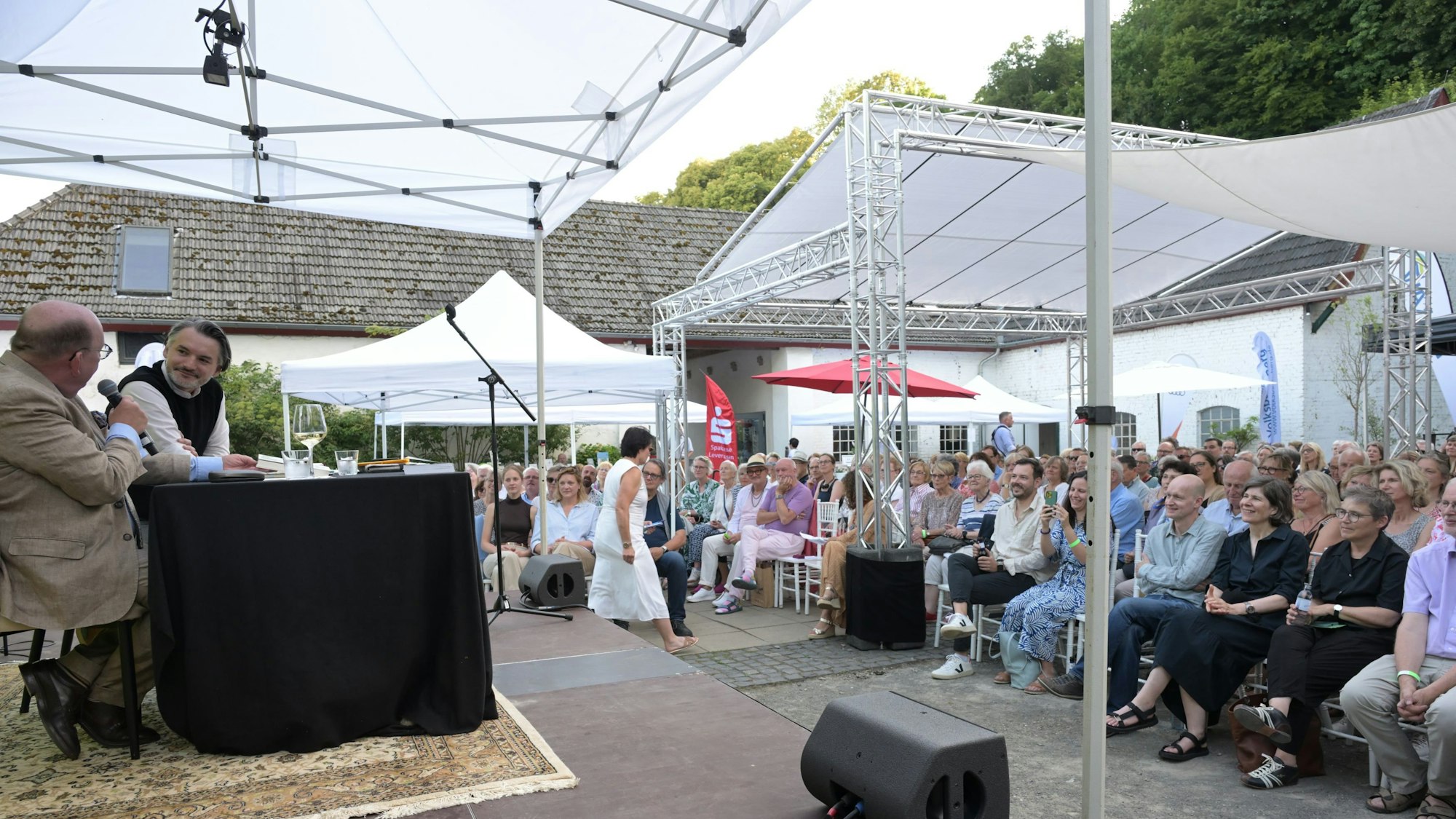 Zwei Männer sitzen auf einer Bühne, davor in einem Innenhof das Publikum unter einem Sonnendach.