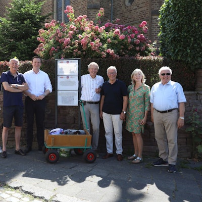 Sieben Männer und eine Frau neben einer Stele mit Info-Tafeln.