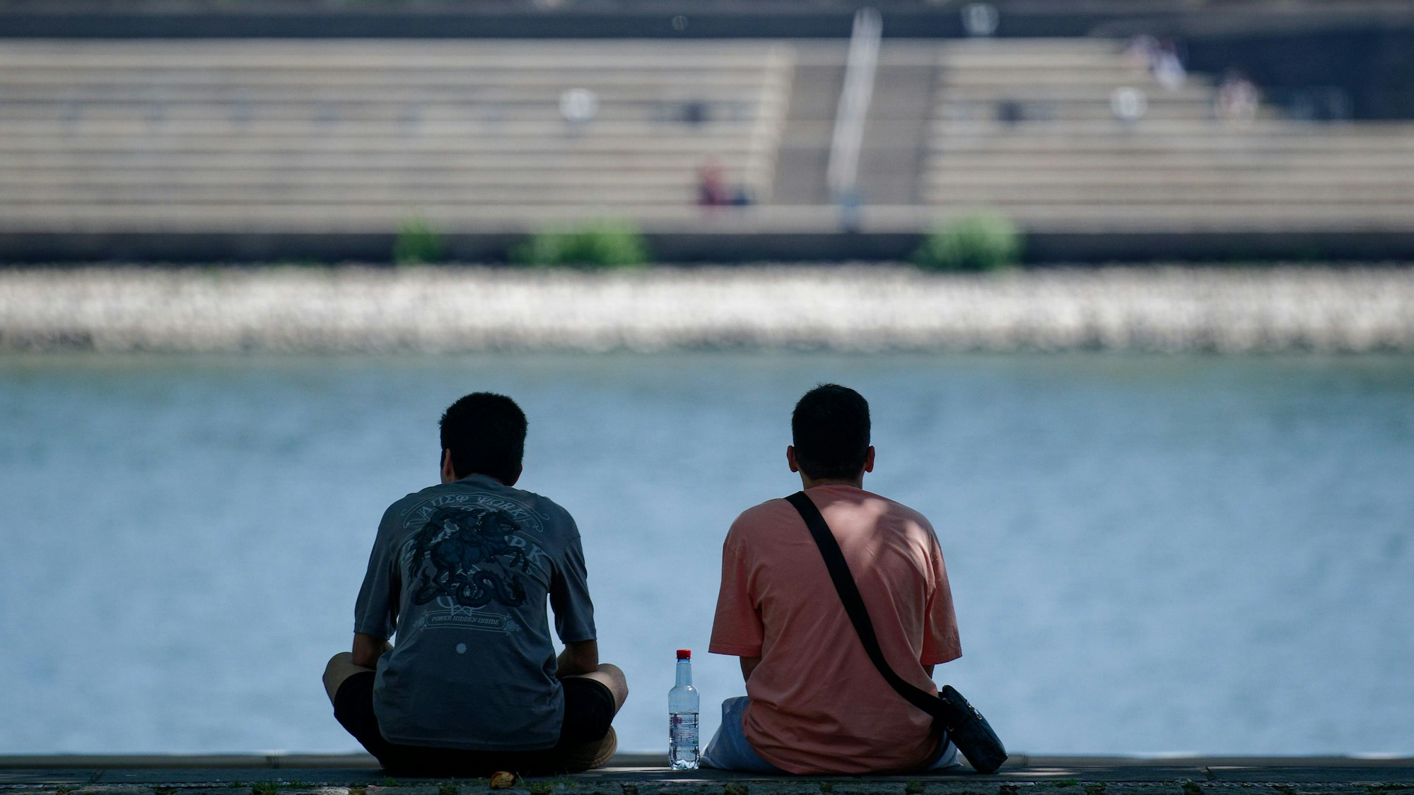 Zwei Männer sitzen mit einer Flasche Wasser am Rhein im Schatten.