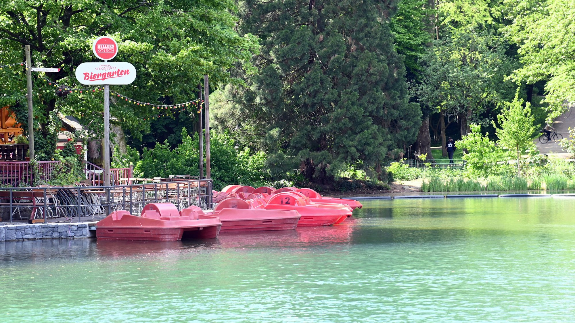 Der Weiher am Volksgarten in der Sonne, neben dem Biergarten liegen Tretboote im Wasser.