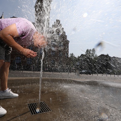Ein Mann hält seinen Kopf in den Wasserstrahl eines Brunnens, der aus dem Boden kommt.
