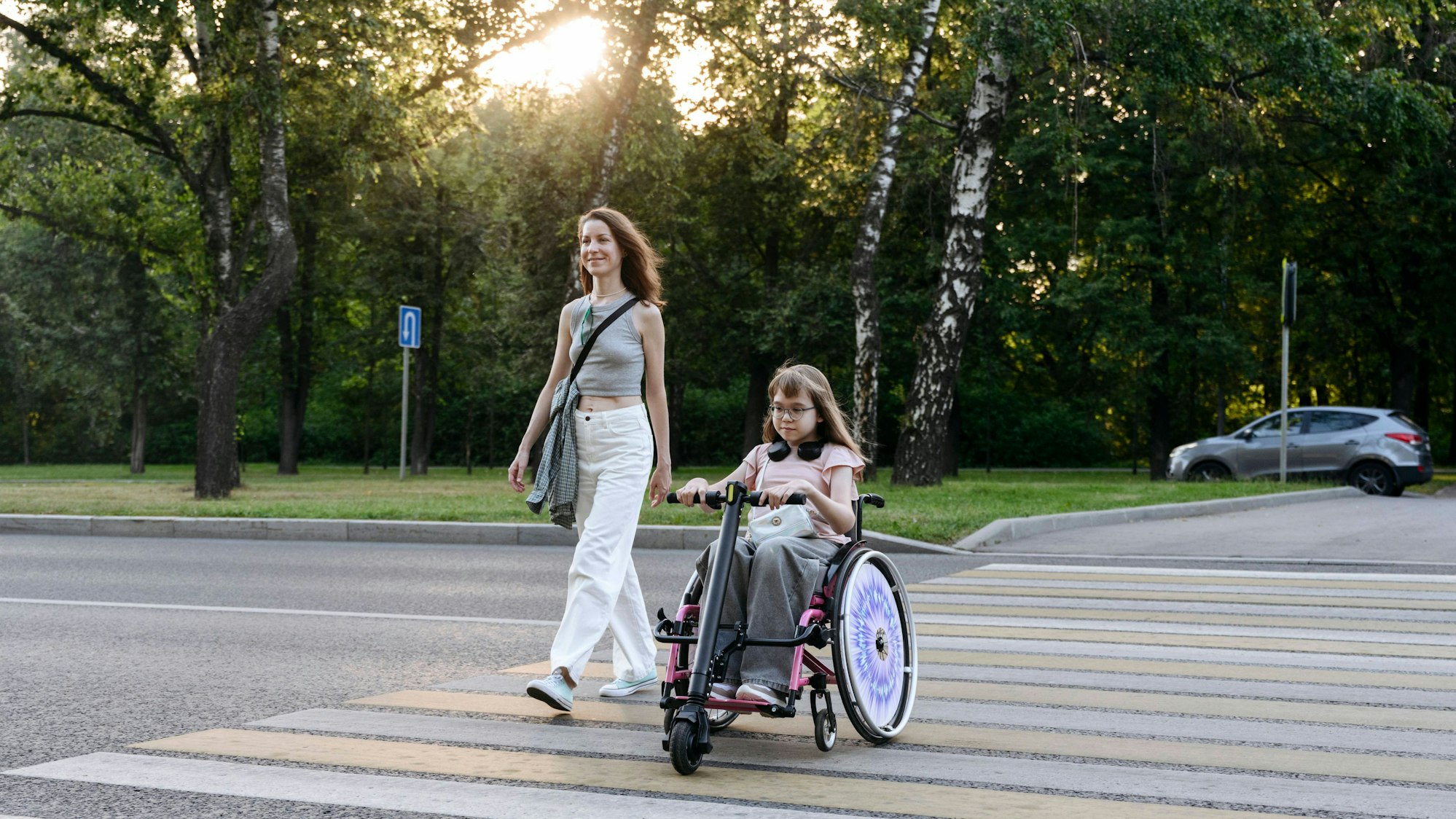 Eine Frau überquert einen Zebrastreifen mit einem Kind, dass in einem Rollstuhl mit Lenker sitzt.