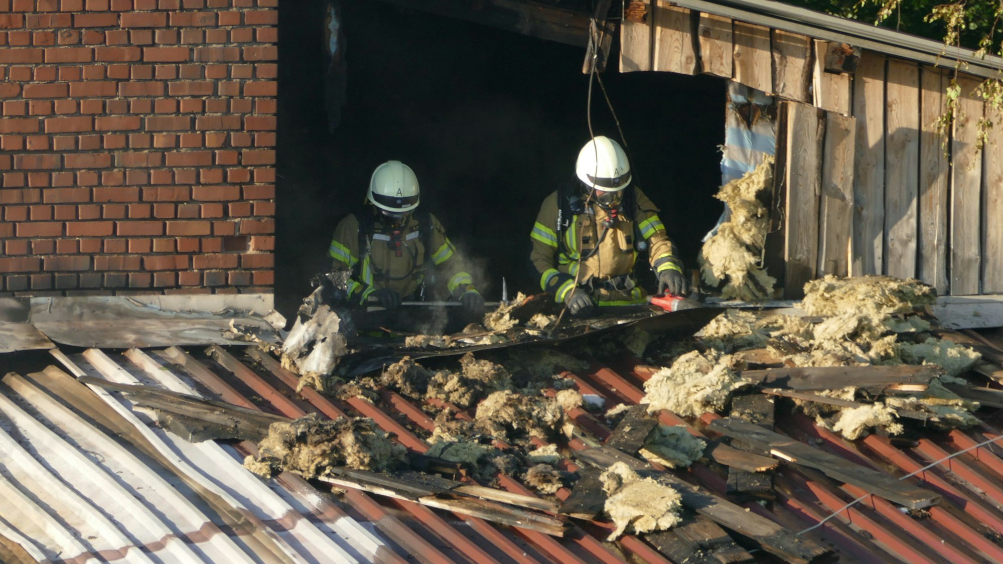 Feuerwehrleute in Atemschutzausrüstung arbeiten in einer Brandruine. Auf dem Dach vor ihnen liegt Dämmwolle.
