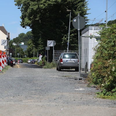 Ein Schotterweg, links stehen rot-weiße Baken, rechts im Hintergrund parkt ein Auto.