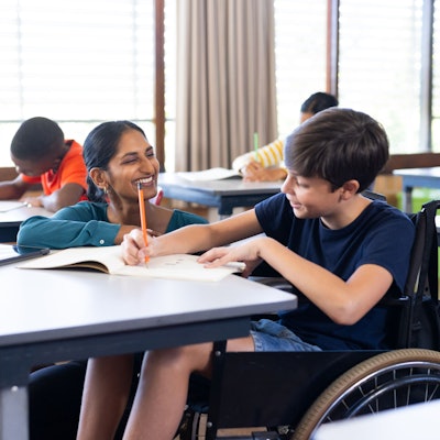Ein Junge sitzt in einem Rollstuhl an der Schulbank und schreibt in ein Heft. Eine Schulbegleiterin kniet neben ihm - und assistiert ihm.
