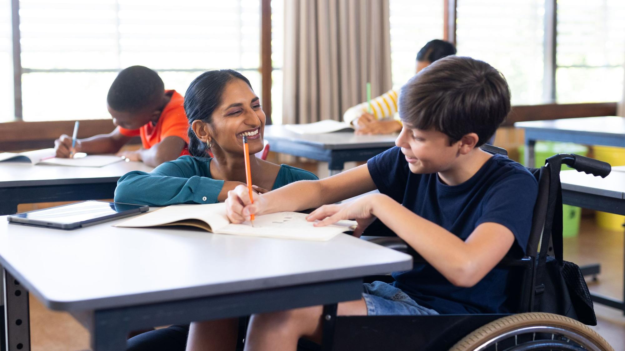 Eine Schulbegleiterin sitzt in einem Klassenraum an dem Tish eines Jungen im Rollstuhl und hilft ihm bei den Schulaufgaben.