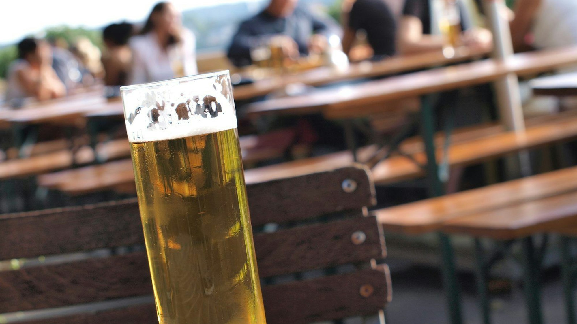 Ein Kölschglas in der Sonne, im Hintergrund sind besetzte Bierbänke zu sehen.