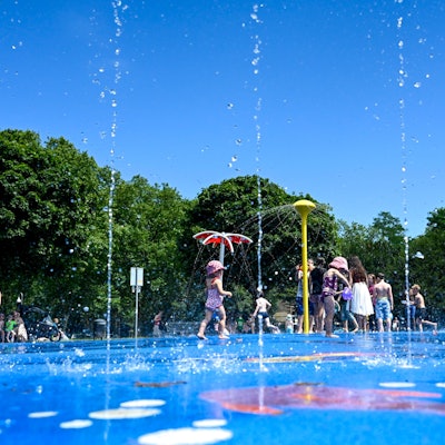 09.07.2024, Köln: Der Wasserspielplatz im Stadtpark ist gut besucht. Kinder spielen im kühlen Nass unter blauem Himmel.