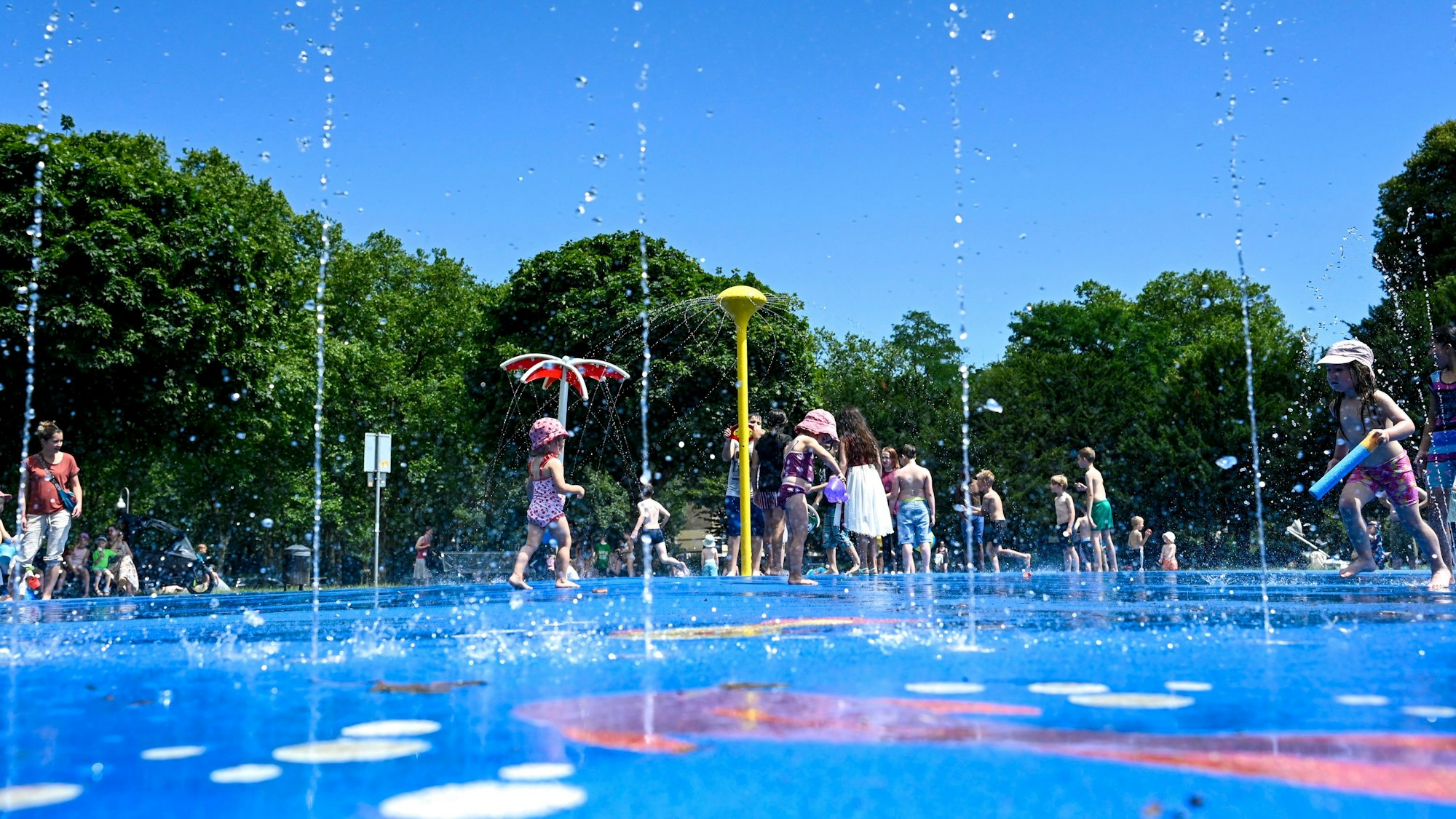 09.07.2024, Köln: Der Wasserspielplatz im Stadtpark ist gut besucht. Kinder spielen im kühlen Nass unter blauem Himmel.