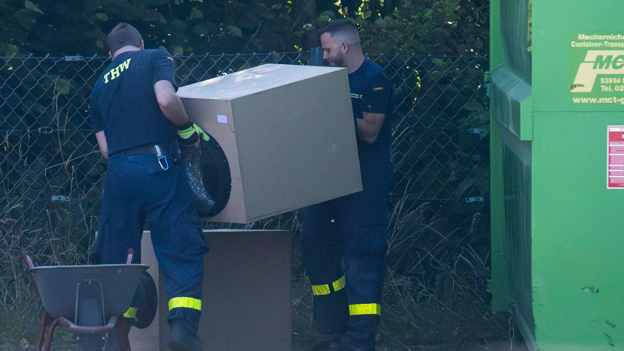 Zwei THW-Mitarbeiter wuchten ein würfelförmiges Bauteil einer Lüftungsanlage aus einer Schubkarre.