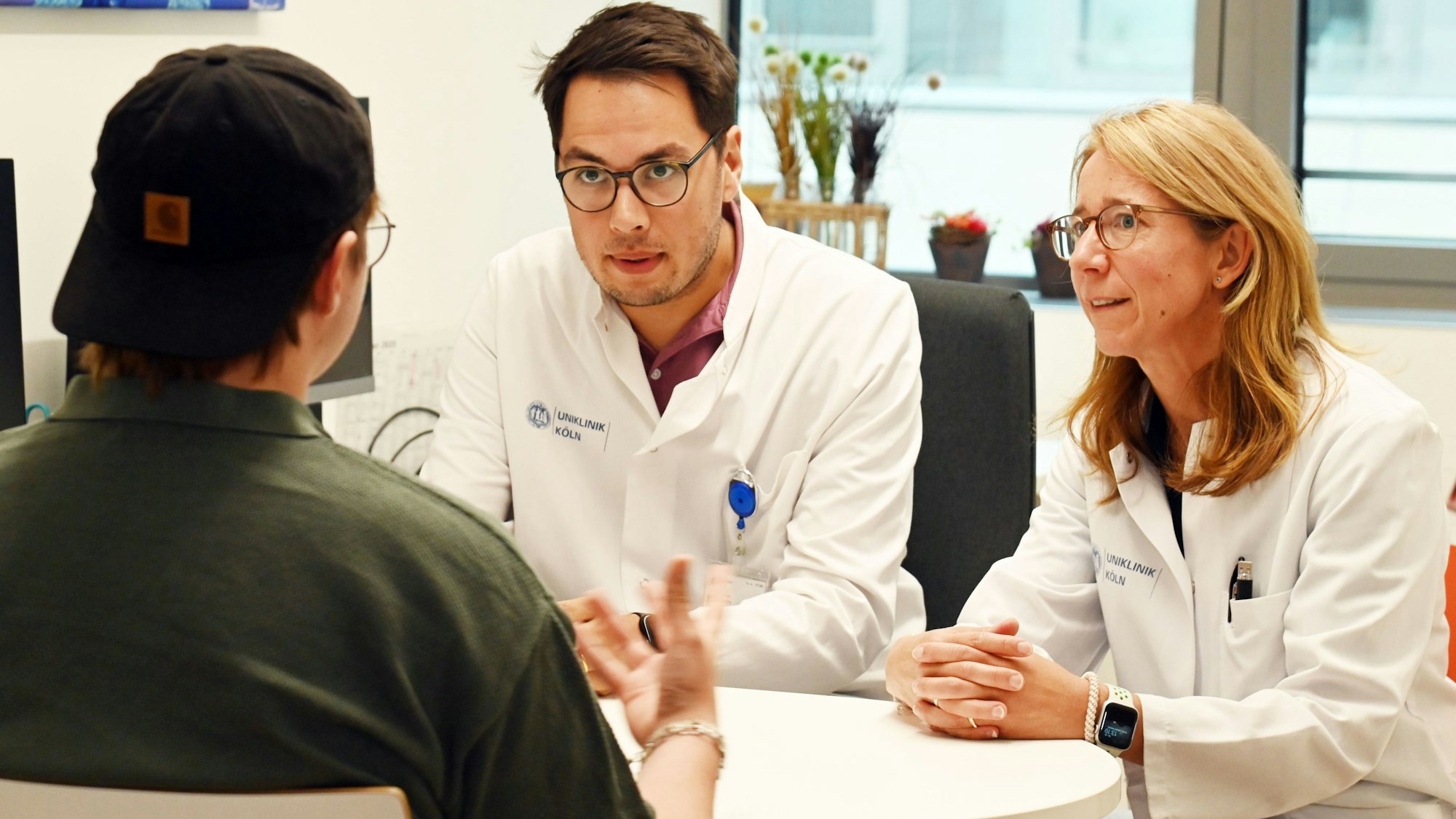 Der Patient ist von hinten zu sehen, er trägt eine schwarze Baseball Cap verkehrt herum auf dem Kopf. Die Ärzte sind mit ihm im Gespräch.