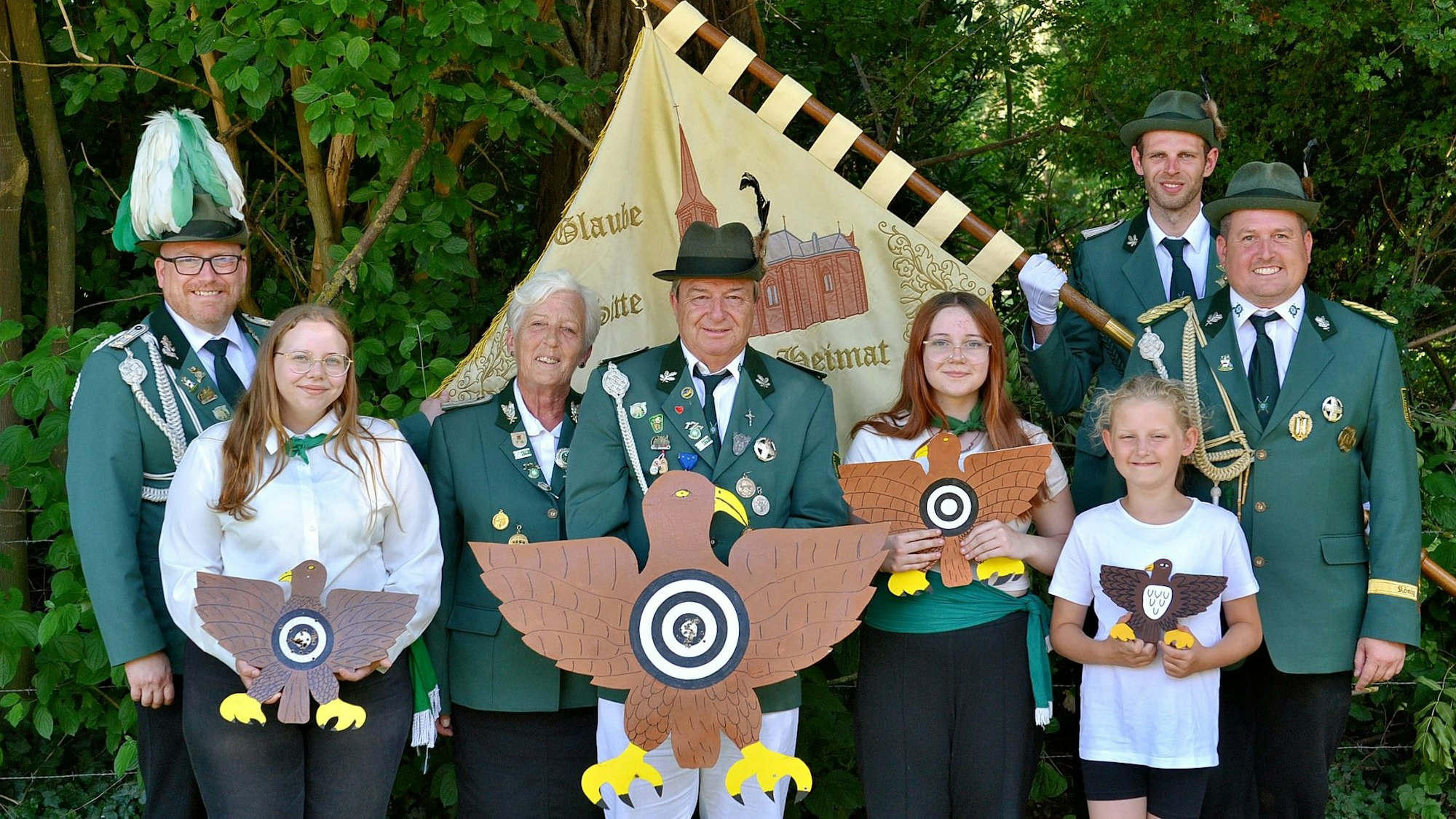 Schützenbrüder und -schwestern stehen mit ihren Trophäen in einer Reihe, hinter ihnen weitere Vereinsmitglieder. Ein Mann hält die Fahne der Bruderschaft.
