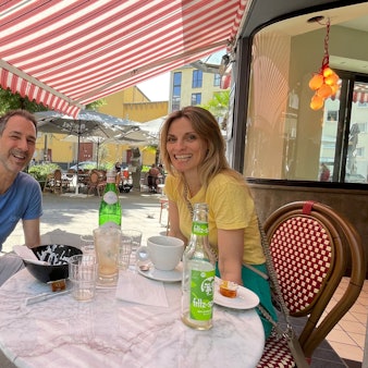 Ein Mann und eine Frau sitzen an einem Café-Tisch draußen, über ihnen spannt sich eine rot-weiße Markise.