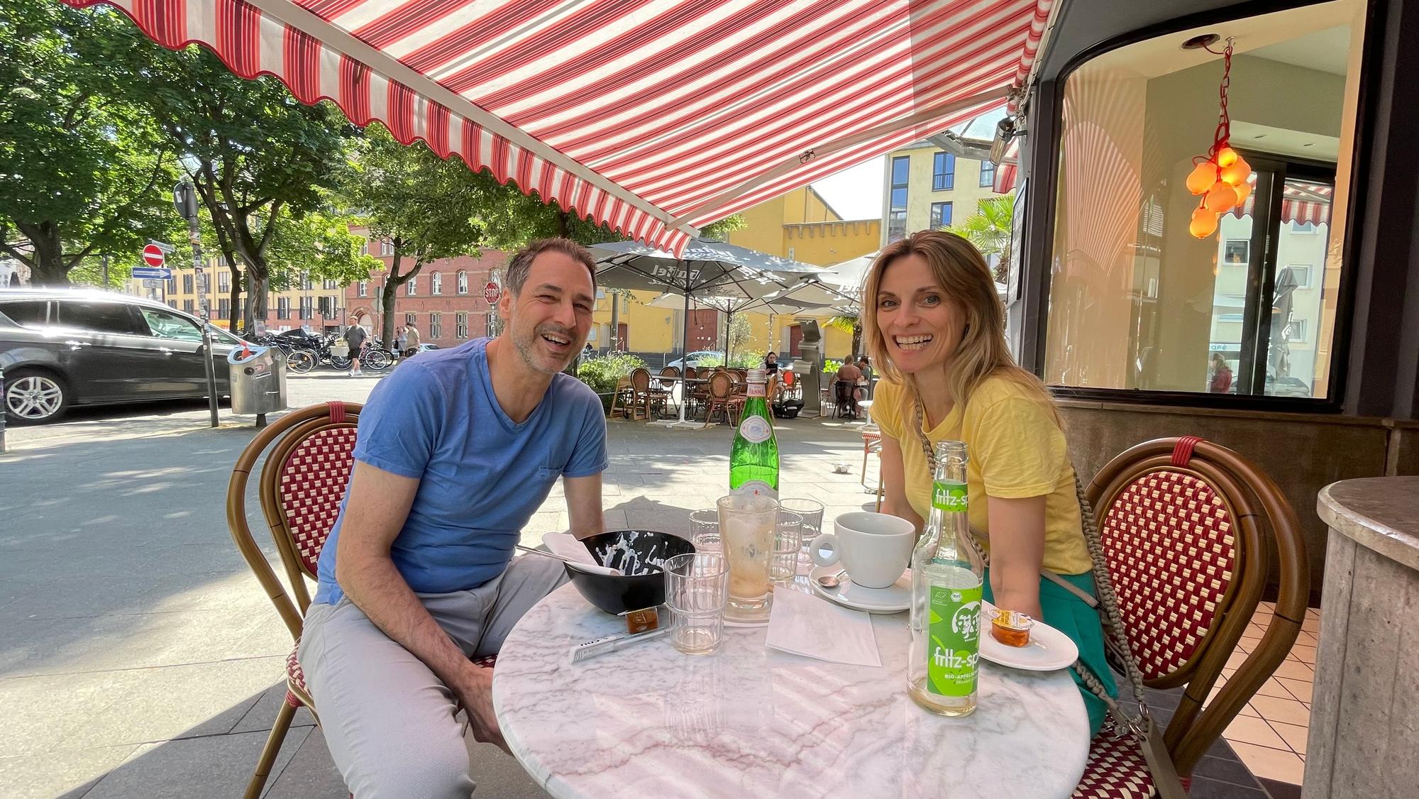 Ein Mann und eine Frau sitzen an einem Café-Tisch draußen, über ihnen spannt sich eine rot-weiße Markise.