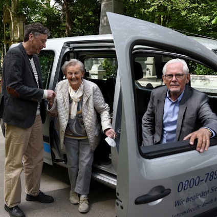 Josef Terfrüchte sitzt im Friedhofsmobil, eine ältere Frau steigt mit der Hilfe eines Mannes aus dem Auto aus.
