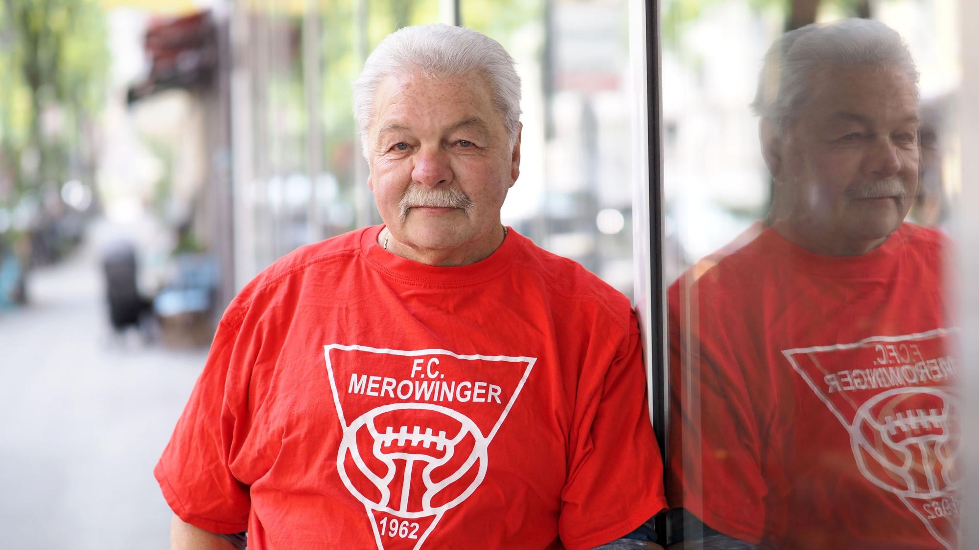 Werner Dive lehnt mit FC-Merowinger-T-Shirt an einer gläsernen Fassade.