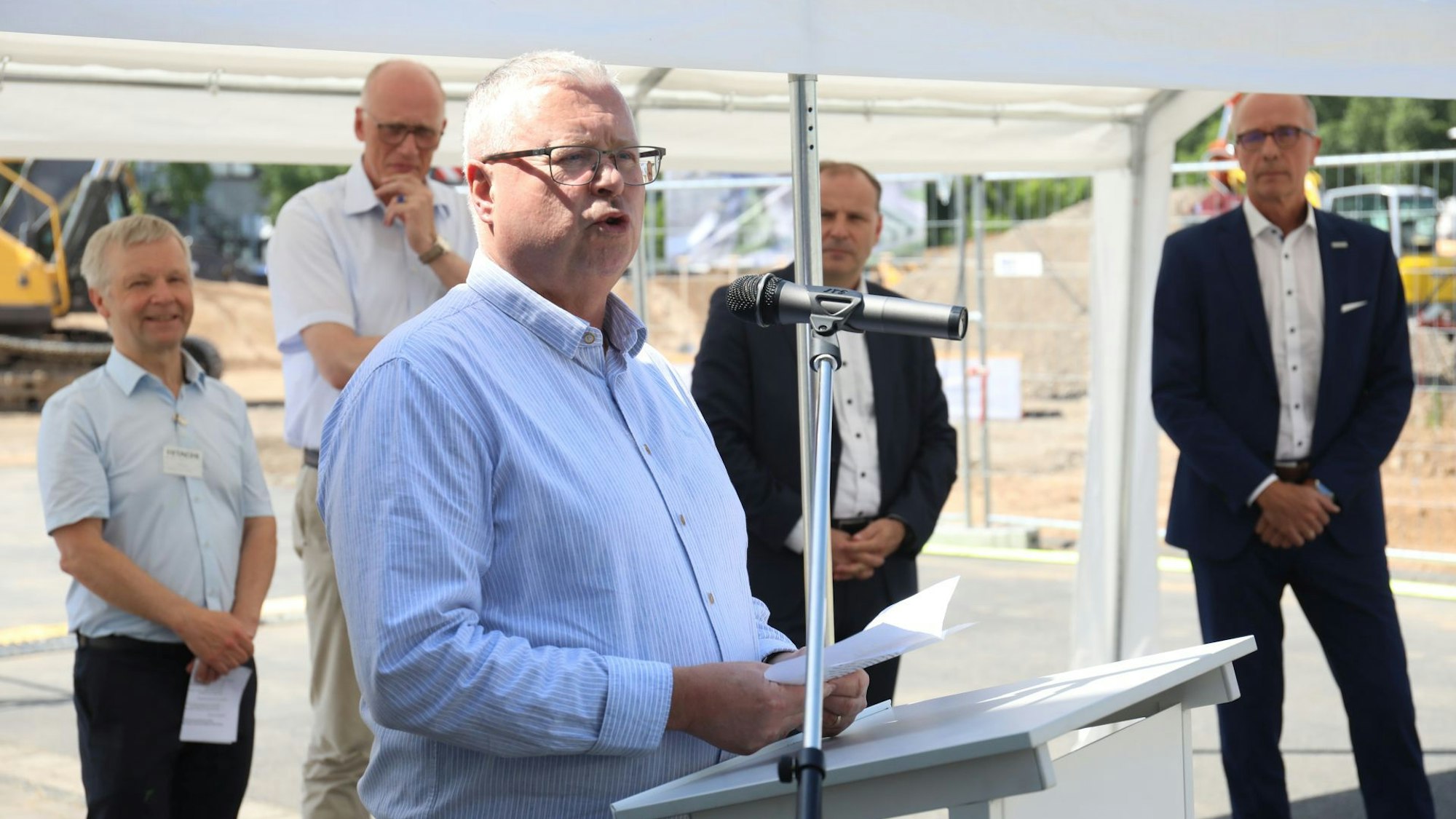 Ein Mann in einem blauen Hemd vor einem Stehpult mit Mikrofon.