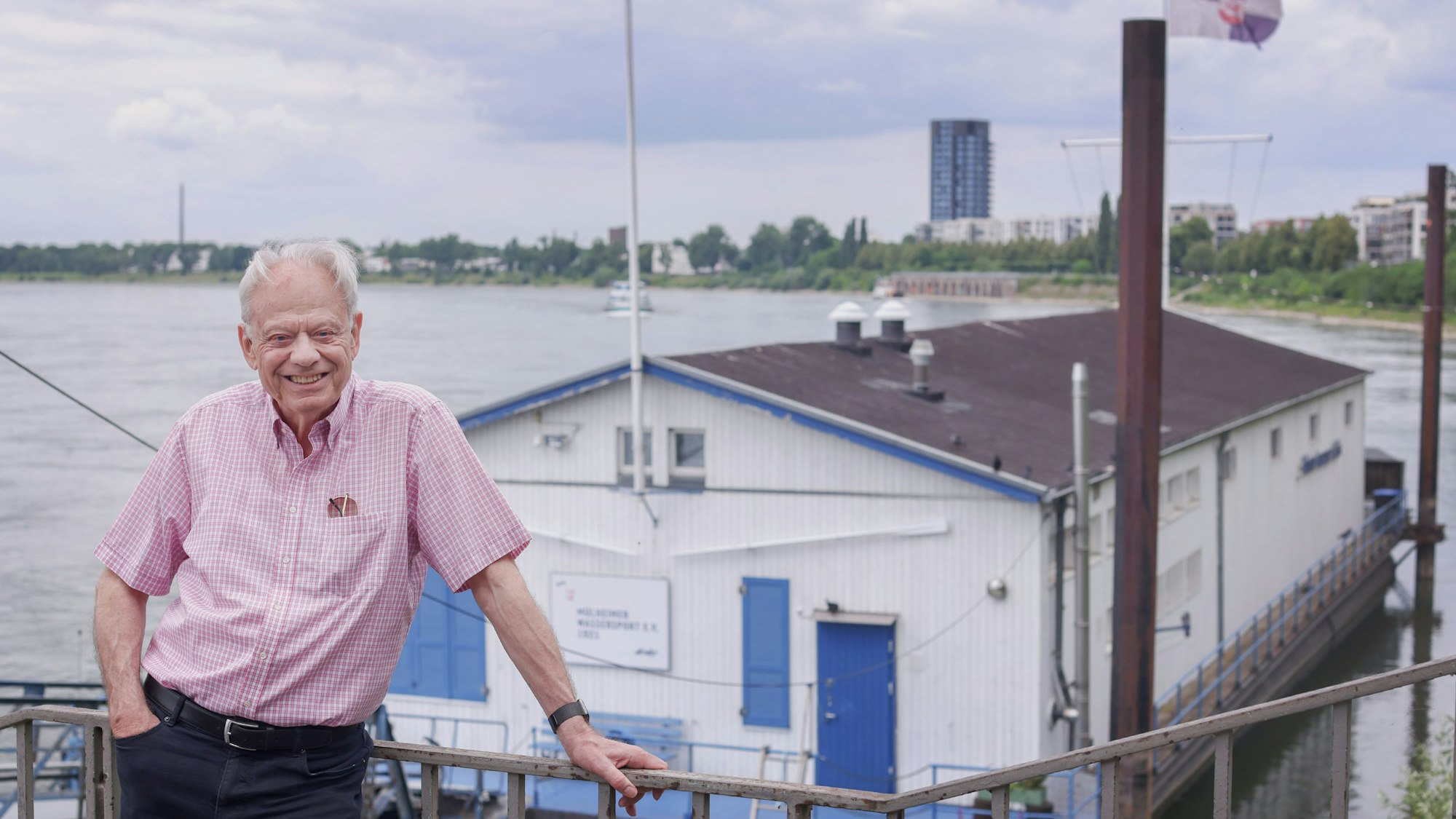 Ein Mann steht an einem Geländer, im Hintergrund auf dem Rhein ist ein großes Bootshaus.