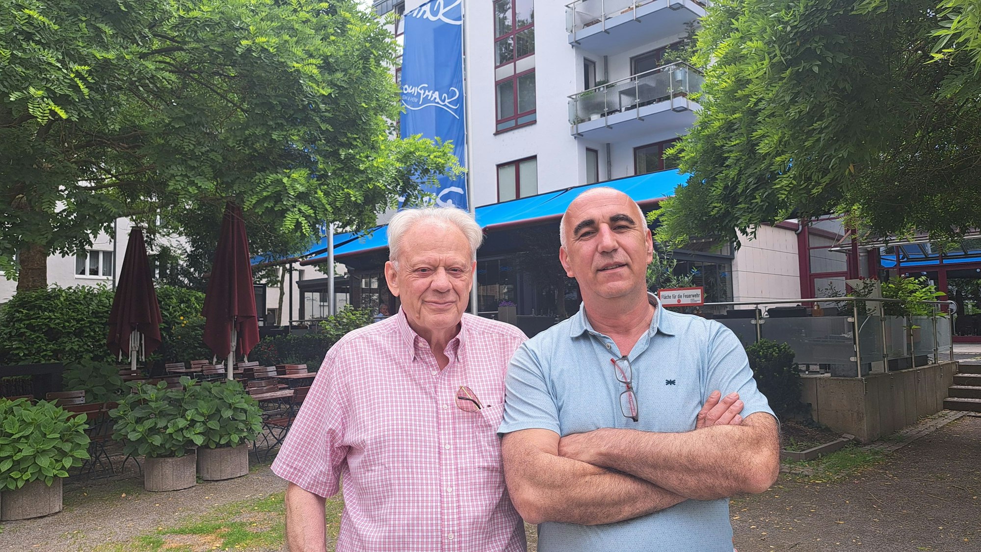 Zwei Männer stehen vor einem mehrgeschossigen Haus, im Hintergrund ist die blaue Markise der Terrasse eines Restaurants zu sehen.