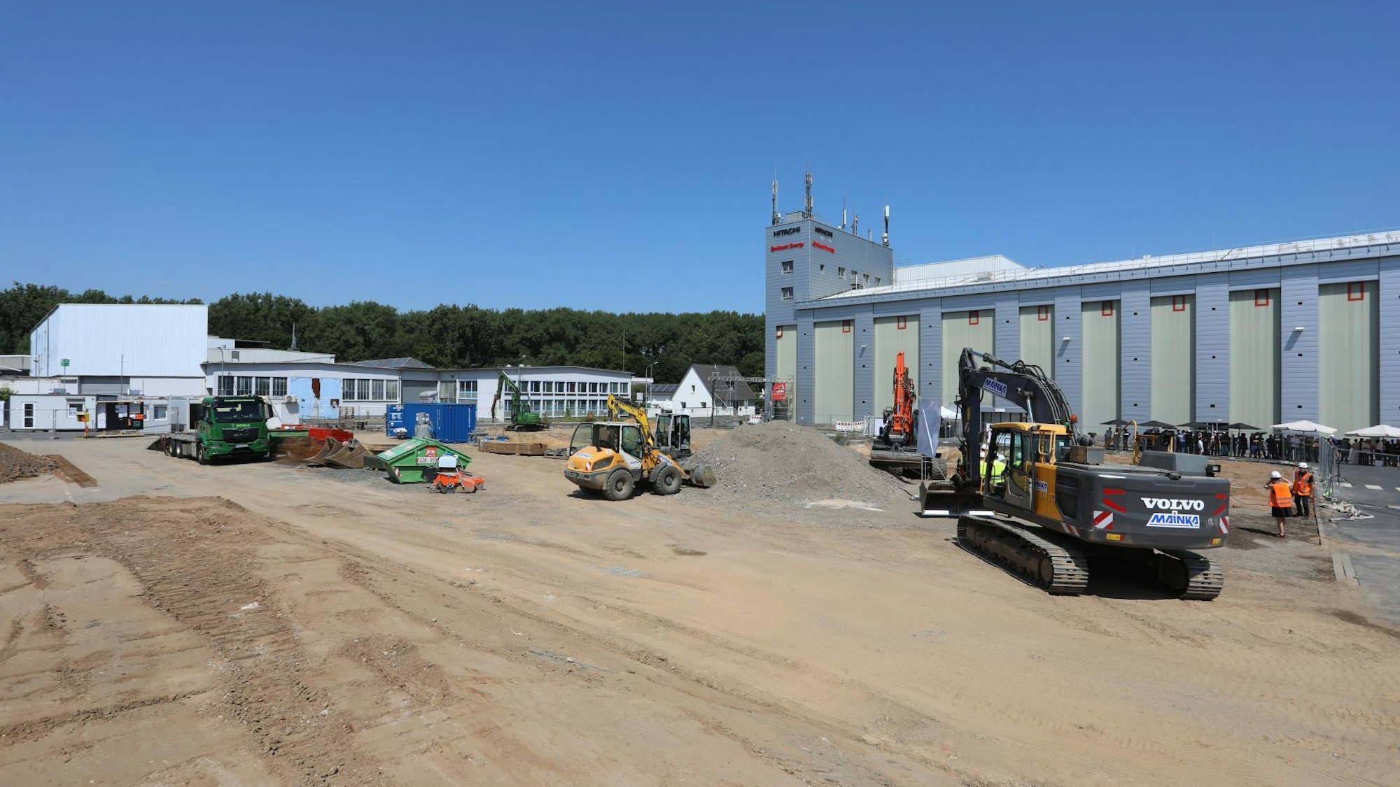 Eine planierte Fläche mit mehreren Baggern, im Hintergrund ein großes Werksgebäude.