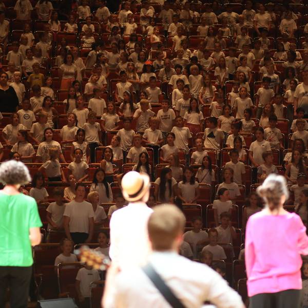 Vier bunt gekleidete Singleiterinnen und Singleiter stehen auf der Bühne der Kölner Philharmonie, die Besucherränge sind bis zum letzten Platz gefüllt mit Grundschulkindern.