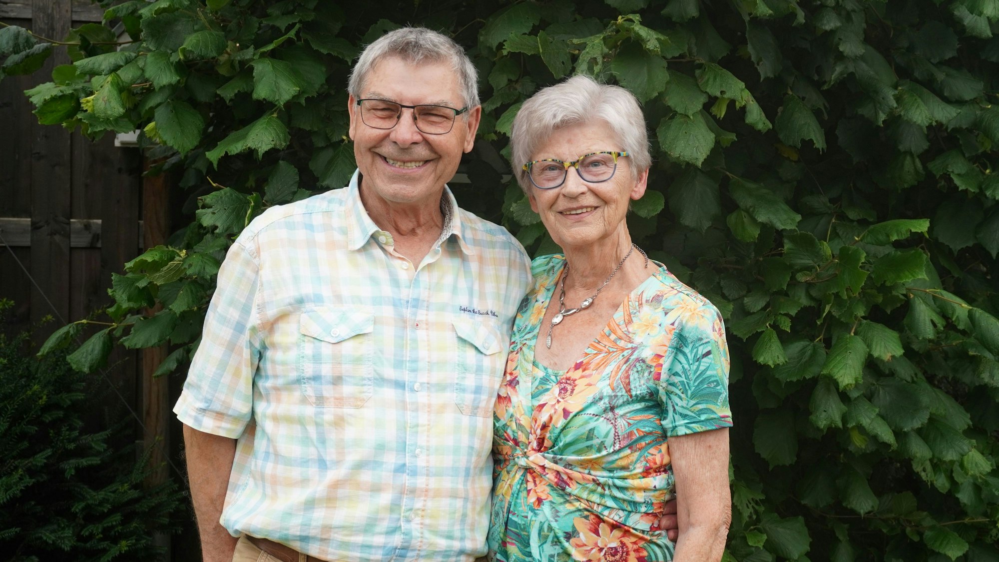 Das Ehepaar steht vor Bäumen und lächelt. Rudi Diekers trägt ein kariertesHemd, Theresia Diekers ein farbenfrohes grün-rotes Oberteil.