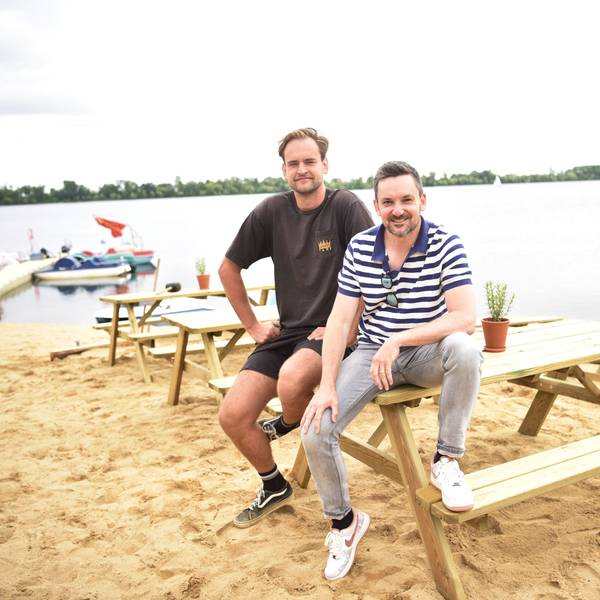 Lukas Funck (links) und Björn Blindert von der surft.GmbH sitzen auf neuen Sitzmöbeln am neuen surft.beach am Zülpicher Wassersportsee. Im Hintergrund sind der See sowie einige Boote zu sehen.