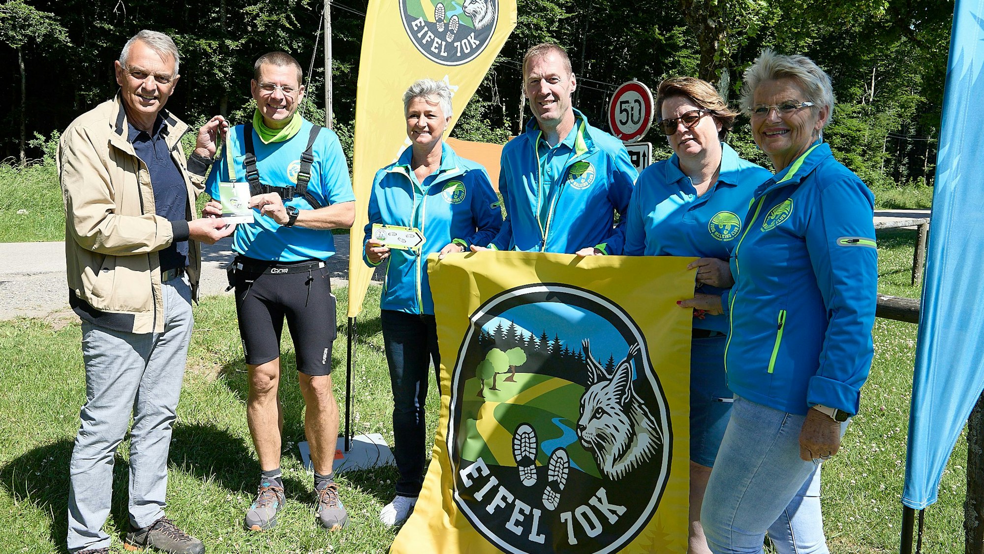 Eine Gruppe steht nahe einem Waldstück bei Bannern, auf denen „Eifel 70K“ steht.